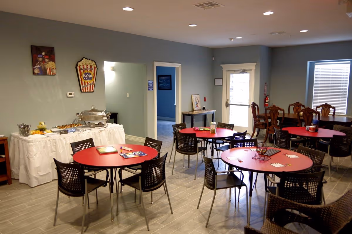 A dining area with several round red tables surrounded by black chairs. A buffet table with a white tablecloth holds food trays and utensils against a light blue wall decorated with popcorn-themed artwork. There is a door and a window with blinds on the right side, and additional wooden chairs and tables are visible in the background.