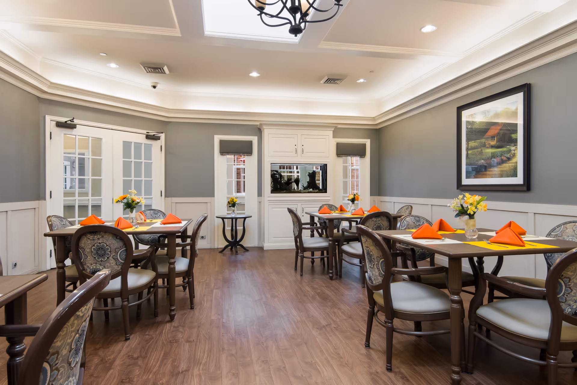 A well-lit dining room with wooden floors and several tables set with orange napkins and small flower arrangements. The walls are painted gray with white wainscoting, and there is a framed painting on the right wall. French doors and windows are visible at the back, along with a built-in cabinet and a fish tank.