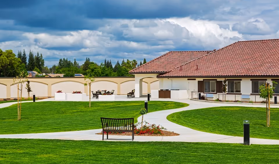 Well-manicured circular lawn and walking paths with a bench and outdoor seating in front of a single-story Mediterranean-style building.