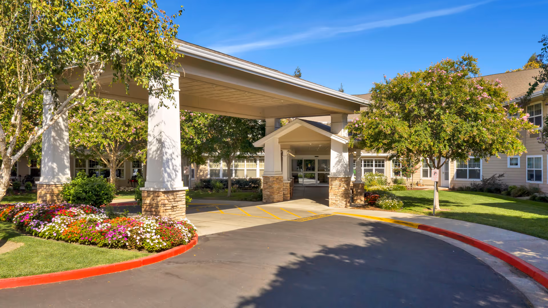 Entrance to a senior living facility with a covered drop-off area supported by white columns with stone bases. The building is surrounded by green lawns, trees, and colorful flower beds under a clear blue sky.