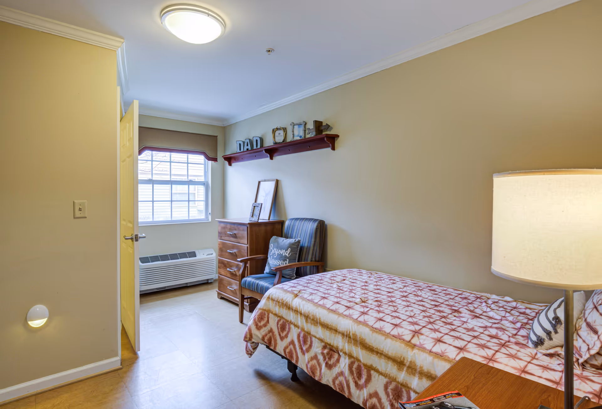 A small bedroom with a single bed covered in a patterned bedspread, a wooden nightstand with a lamp, a striped armchair with a pillow that says 'Beyond Blessed', a wooden dresser, and a window with a beige valance. A shelf above the dresser holds decorative items including letters spelling 'DAD'. The walls are painted light beige and the floor is a light wood or laminate.