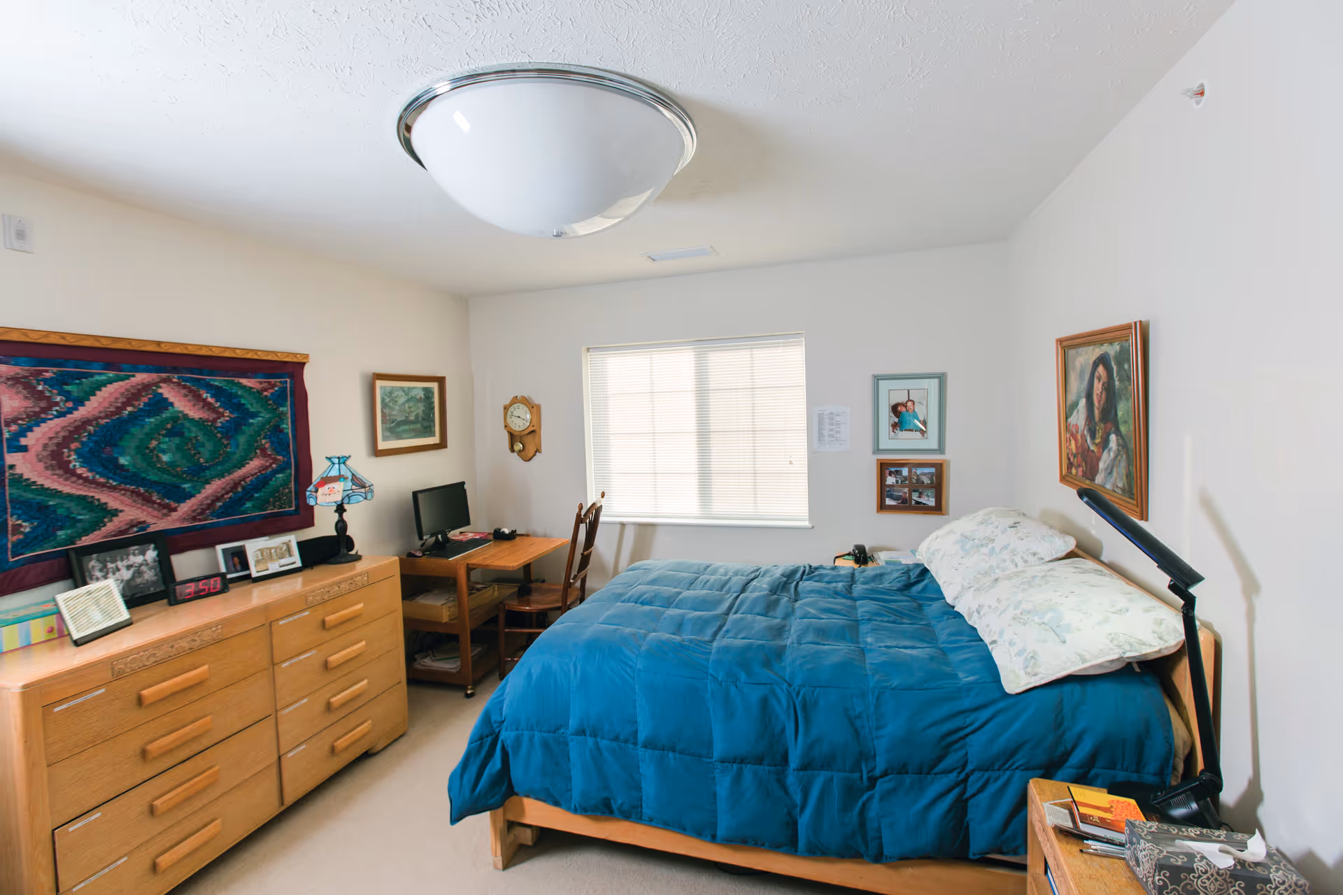 A cozy bedroom with a wooden bed frame and a blue comforter. There are two pillows with floral pillowcases on the bed. A wooden dresser with several framed photos and a digital clock is on the left side of the room. A small wooden desk with a computer monitor and chair is positioned near a window with closed blinds. The walls are decorated with various framed pictures and a colorful woven wall hanging. A ceiling light fixture is visible above.