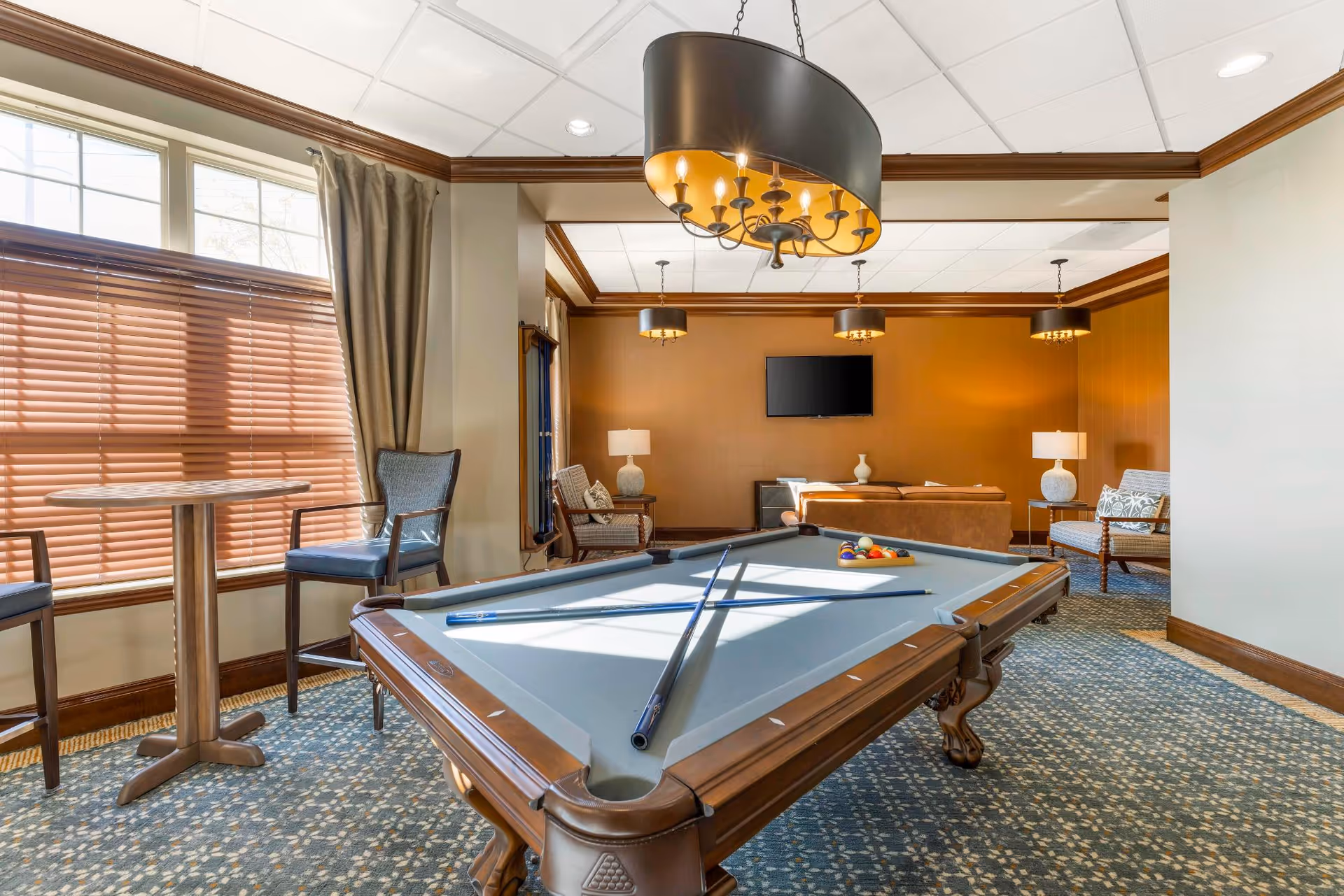 A well-lit recreational room featuring a pool table with cues and balls arranged on it. The room has patterned carpet flooring, large windows with blinds and curtains, a small round table with chairs, and a seating area with armchairs and a sofa facing a wall-mounted flat-screen TV. Warm lighting fixtures hang from the ceiling, and the walls are painted in neutral tones with wood trim accents.