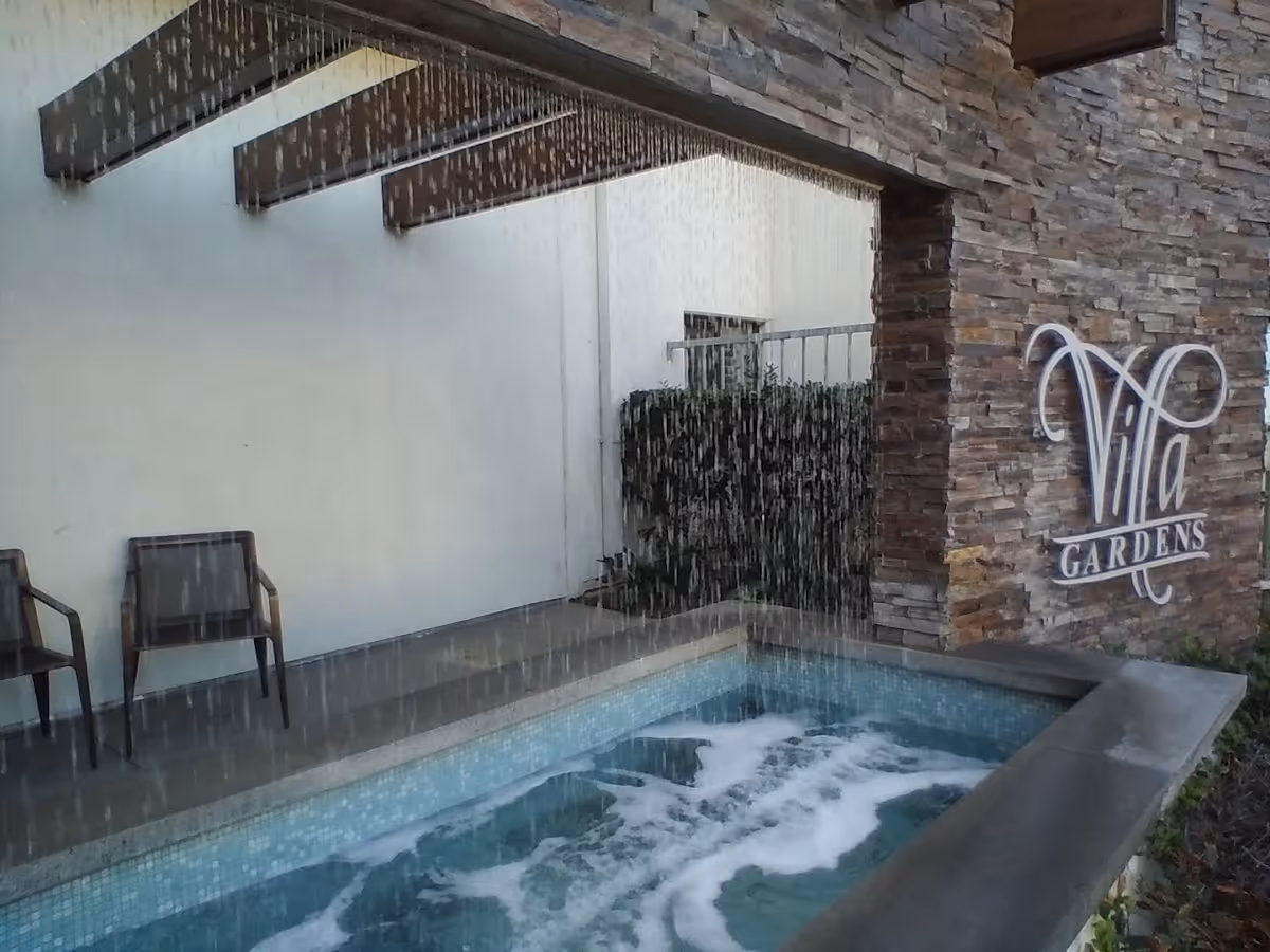 Outdoor water feature with a small pool and water cascading down from a wooden pergola structure. Two chairs are placed on the patio beside the pool. A stone wall with the sign 'Villa Gardens' is visible in the background.