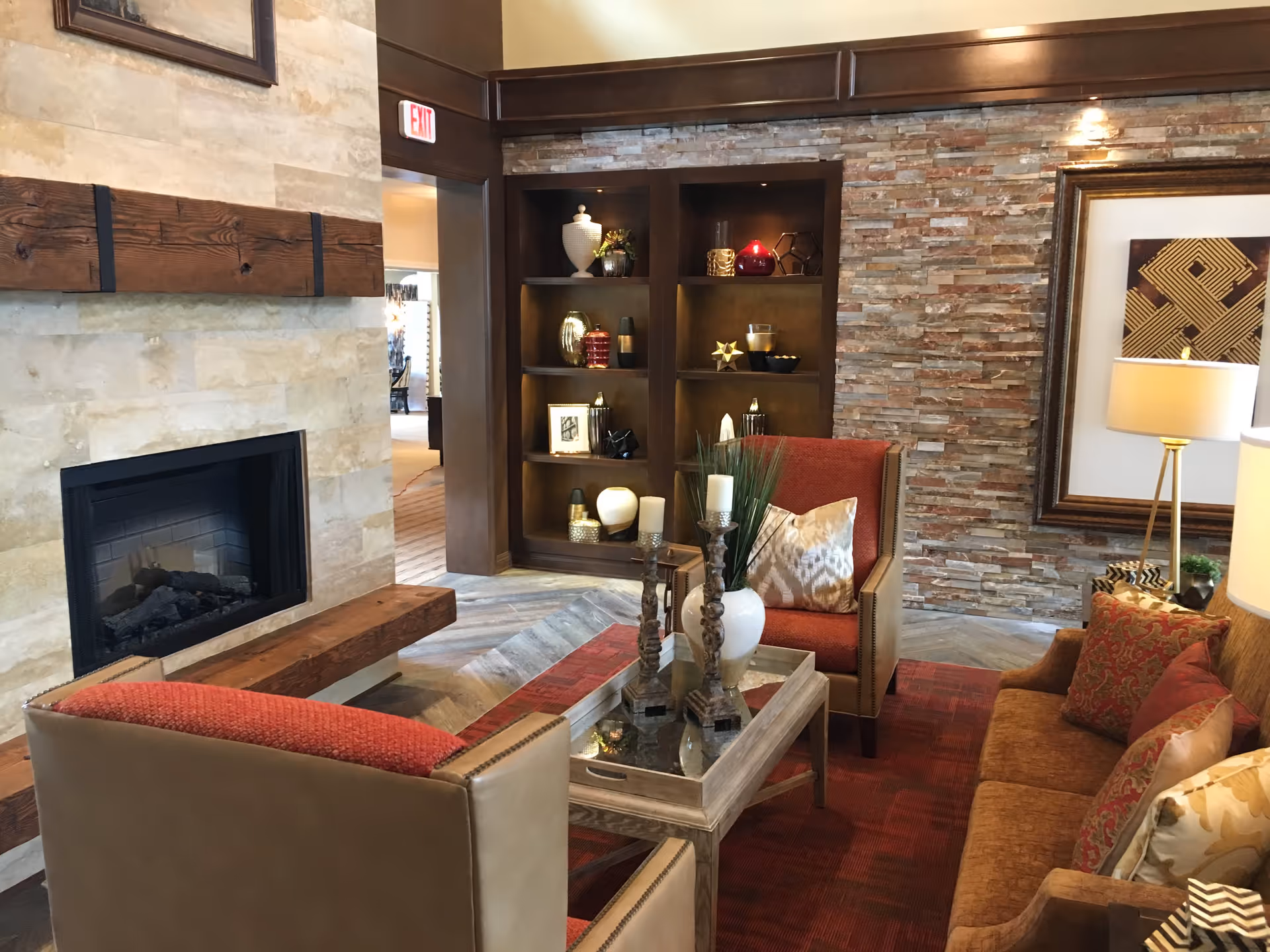 Cozy living room area with a stone fireplace on the left, wooden mantel, and built-in shelves decorated with vases and ornaments. There are two armchairs with red cushions and a brown sofa with patterned pillows around a glass-top coffee table holding candles and a vase with greenery. The walls feature stone and wood paneling, and a framed abstract artwork is visible on the right wall next to a floor lamp.