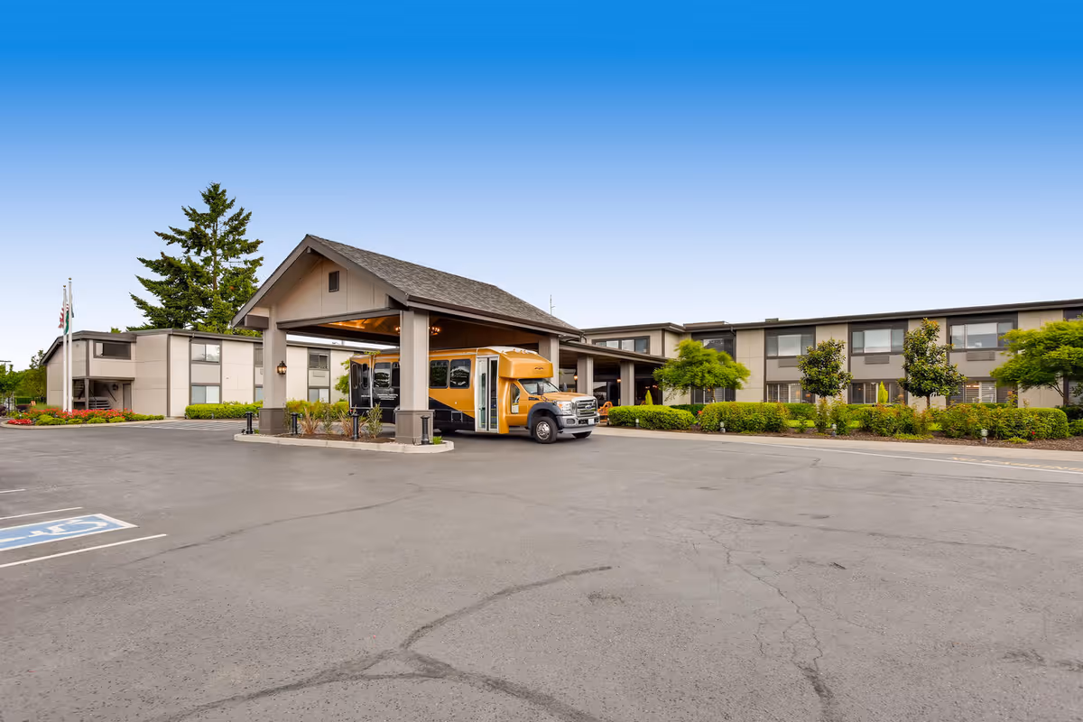 Exterior view of Cascade Inn Assisted Living Community showing a two-story building with a covered entrance where a yellow shuttle bus is parked. The area is surrounded by greenery and trees under a clear blue sky.