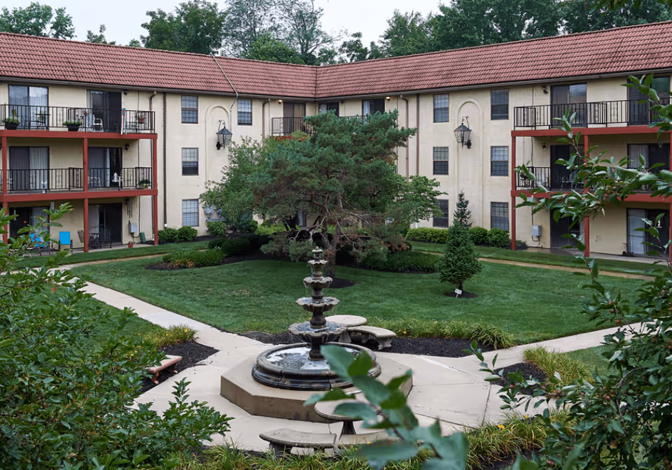 Outdoor courtyard area of a senior living facility with a central multi-tiered water fountain surrounded by green grass, trees, and shrubs. The courtyard is enclosed by a three-story building with balconies and windows, and paved walkways lead to the fountain.