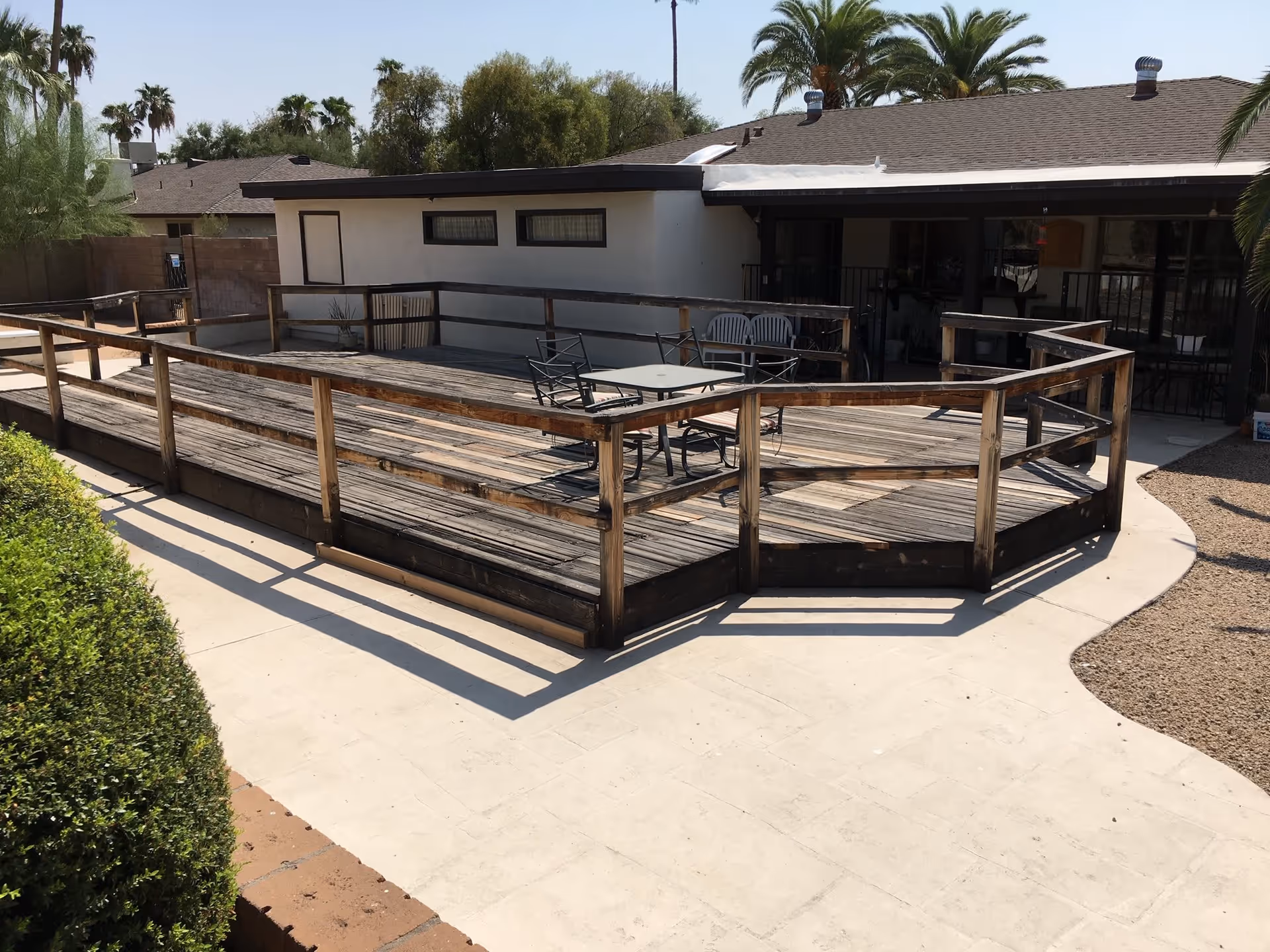 Outdoor wooden deck with a railing surrounding it, featuring a table and chairs. The deck is adjacent to a single-story building with a shaded patio area. Palm trees and other greenery are visible in the background under a clear sky.