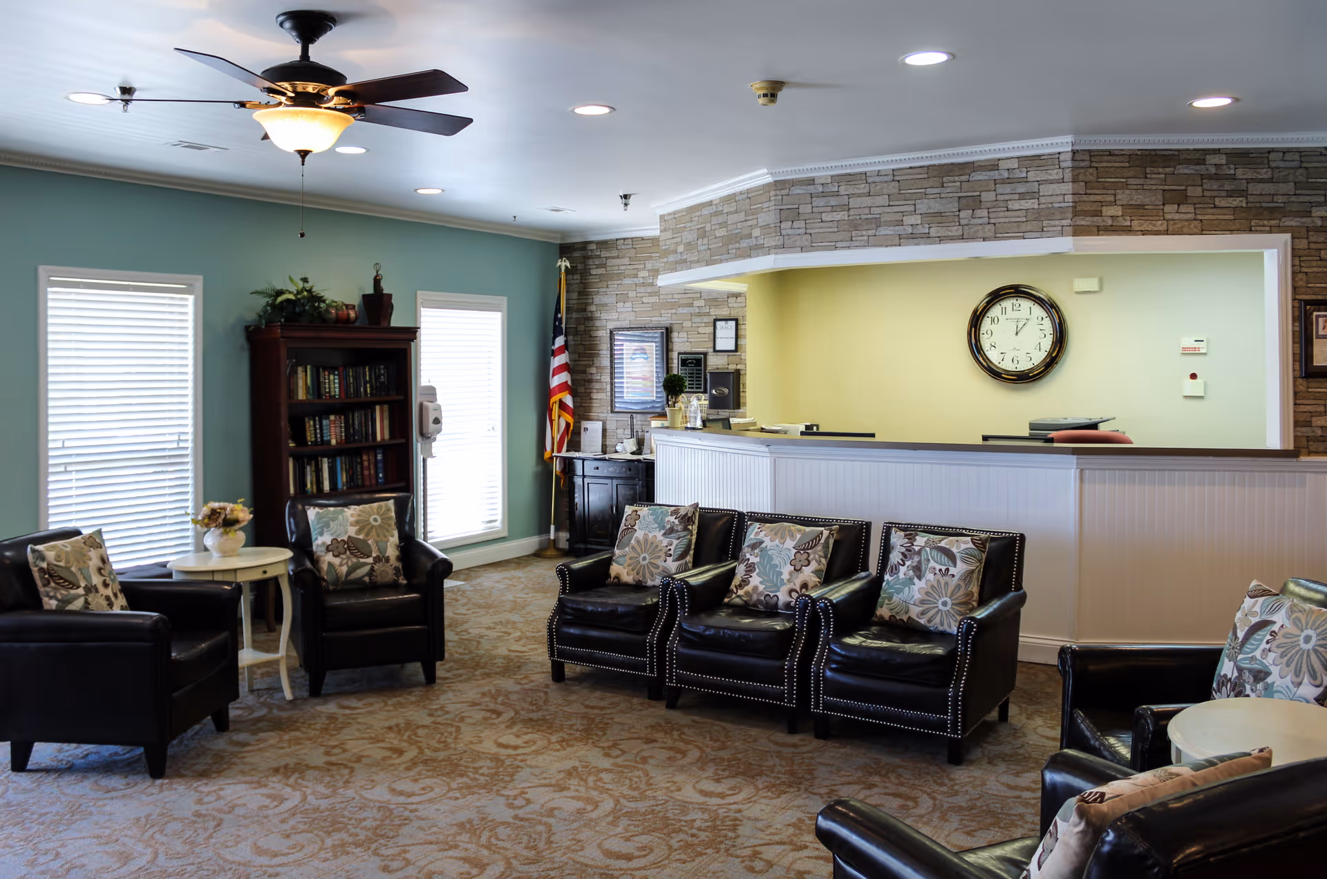 A cozy seating area in a senior living facility with six black leather armchairs adorned with floral patterned cushions arranged around small white tables. The room features a beige patterned carpet, light blue walls with two windows covered by white blinds, a wooden bookshelf filled with books, and an American flag in the corner. A reception desk with a stone accent wall behind it and a large clock is visible in the background. The ceiling has recessed lighting and a ceiling fan with a light fixture.