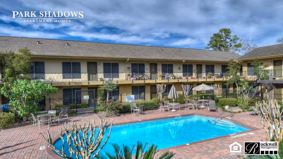 Outdoor view of Park Shadows Apartment Homes featuring a rectangular swimming pool surrounded by a brick patio with tables, chairs, and umbrellas. The two-story apartment building with balconies and windows is visible in the background under a partly cloudy sky.
