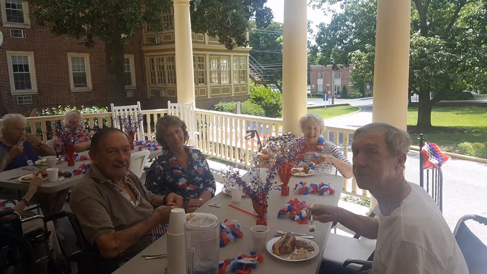 A group of elderly people sitting around a table on a covered outdoor patio, enjoying a meal together. The table is decorated with red, white, and blue patriotic decorations, and there are trees and buildings visible in the background.