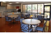 A dining area with round tables and blue cushioned chairs arranged around them. The room has large windows and a glass door letting in natural light, with a kitchen area visible in the background featuring appliances and cabinetry.