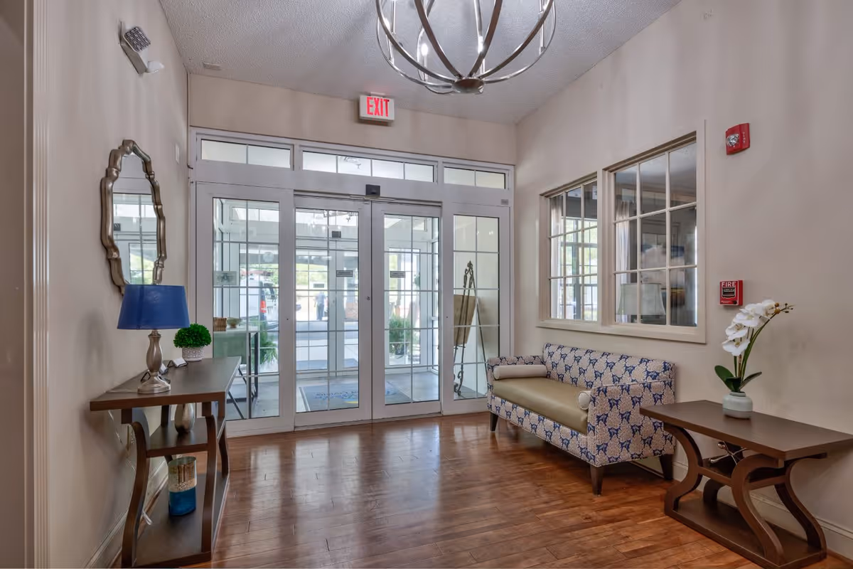 Entrance area of a facility with glass double doors and an exit sign above. Inside, there is a patterned couch with a bolster pillow on the right, a wooden side table with a white orchid plant, and another wooden console table on the left with a blue lamp and a small green plant. A decorative mirror hangs above the console table, and a modern chandelier is visible on the ceiling. The floor is wooden, and there are large windows on the right wall.