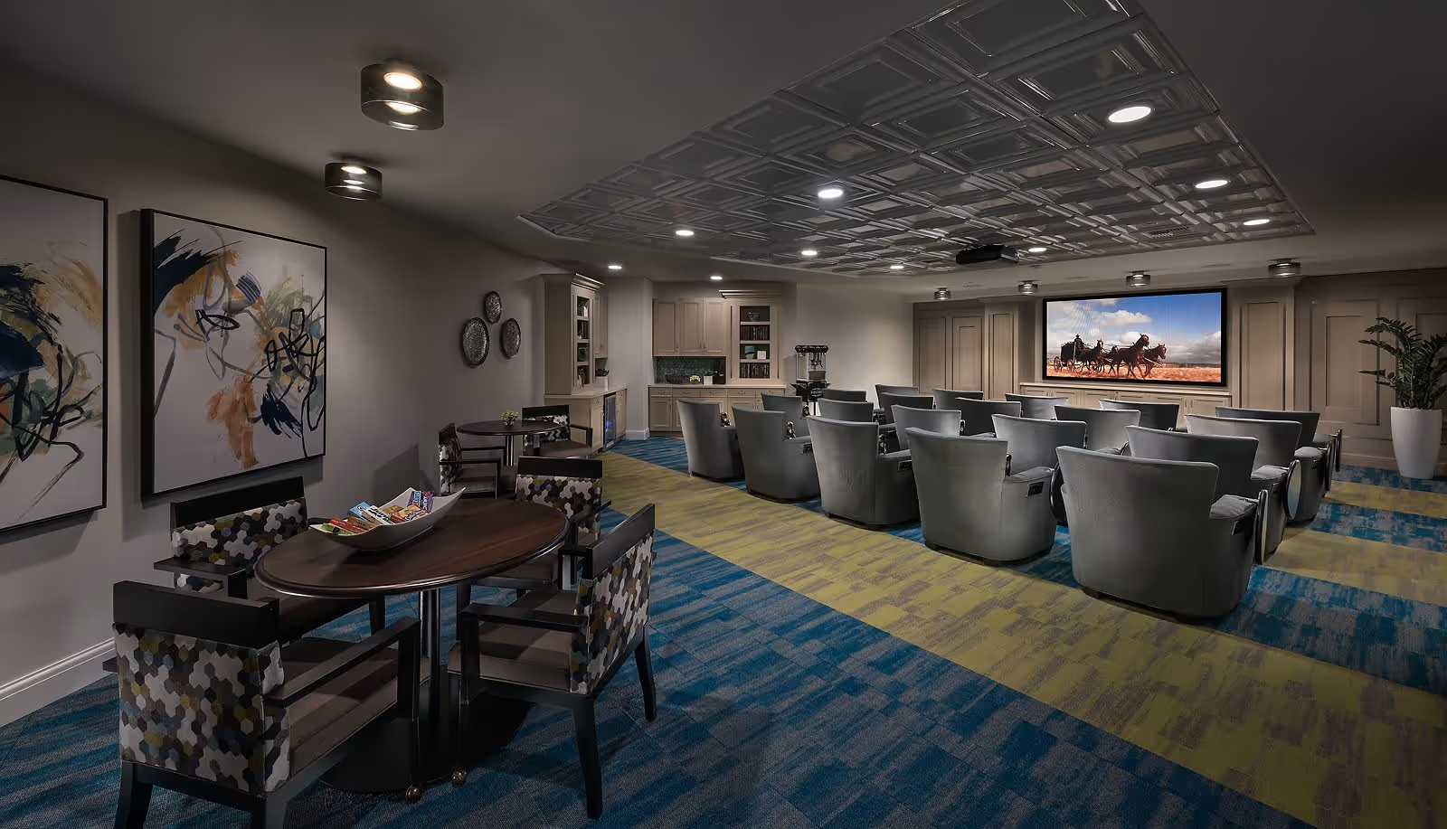 A cozy media room with multiple gray armchairs arranged in rows facing a large screen showing a western scene with horses. The room features a patterned blue and green carpet, a ceiling with recessed lighting, and a small round table with four patterned chairs on the left side. Abstract artwork and decorative plates adorn the walls, and a potted plant is placed in the corner.