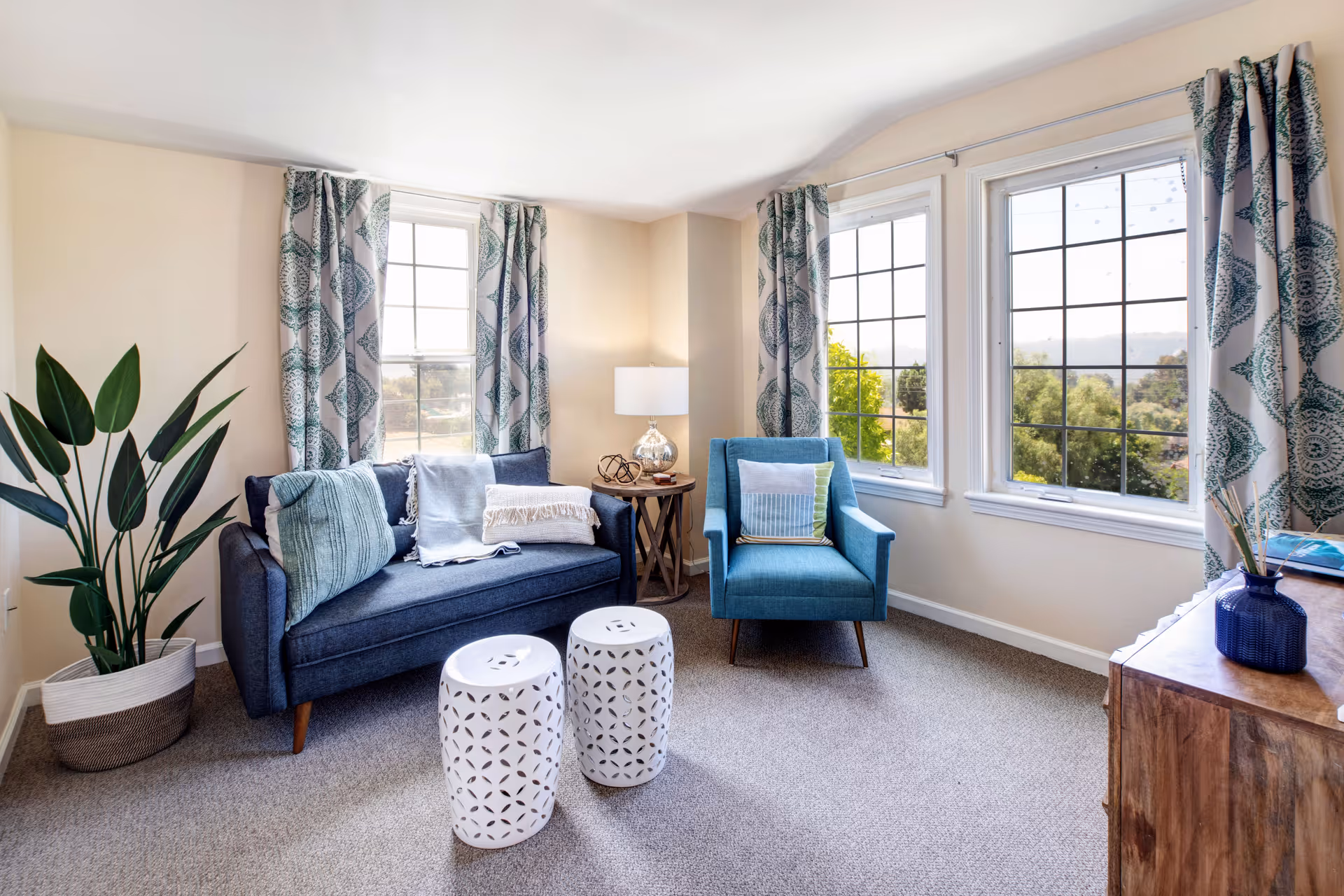 A bright living room with large windows covered by patterned curtains, a dark blue sofa with cushions and a throw blanket, a blue armchair with a cushion, two white decorative stools, a wooden side table with a lamp and decorative items, a large potted plant in the corner, and a wooden cabinet with a blue vase on top.