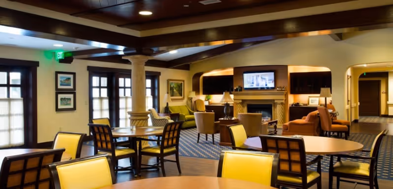Spacious senior living common room with round dining tables, yellow chairs, lounge seating, fireplace and a TV.