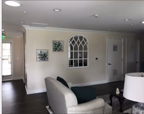 Interior view of a living room area in a senior living facility with a striped sofa, dark green cushions, a coffee table with decorative items, a table lamp, two framed botanical artworks on the wall, and a decorative arched window frame. There are doors and a hallway visible in the background.