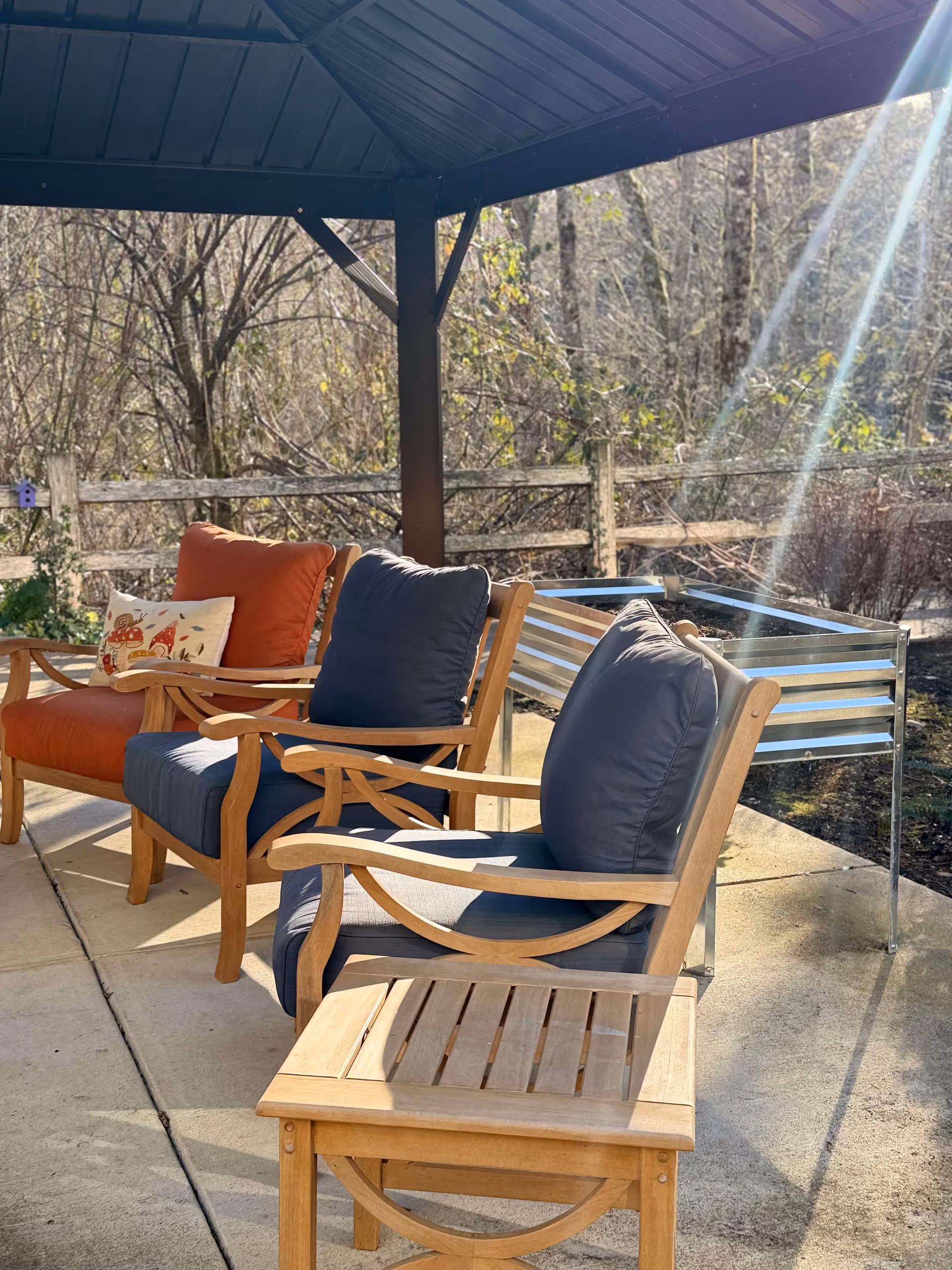 Outdoor seating area under a covered patio with wooden chairs featuring blue and orange cushions, a small wooden table, and a metal planter box. Sunlight streams through the trees and a wooden fence is visible in the background.
