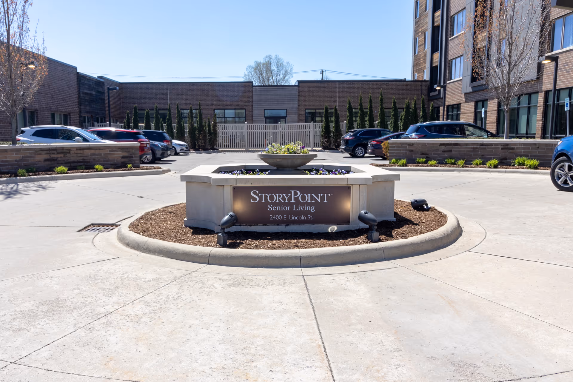 Entrance area of StoryPoint Senior Living facility showing a circular driveway with a central planter that has flowers and a sign displaying the facility name and address. Several parked cars are visible on either side, with the building and some trees in the background under a clear blue sky.