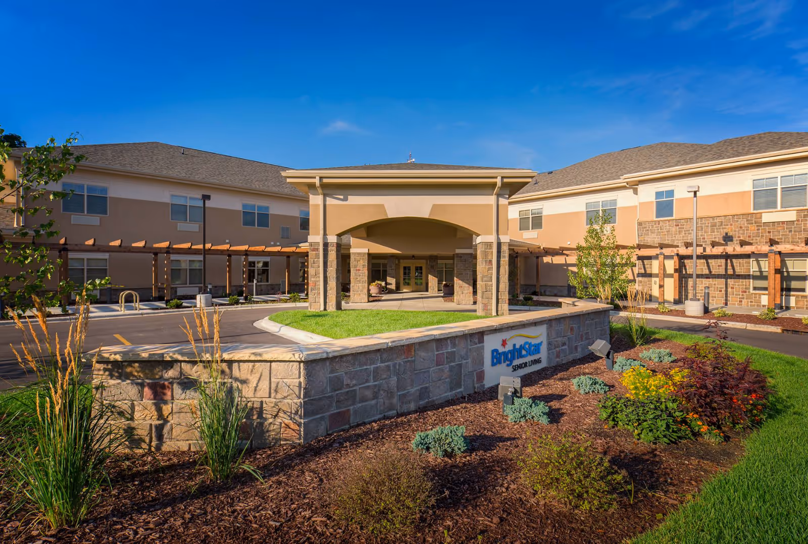 Exterior view of Brightstar Senior Living facility showing a two-story building with a covered entrance, landscaped garden with various plants, and a stone wall with the facility's sign.