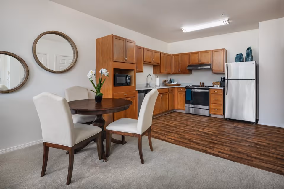 Open kitchen with wooden cabinets and stainless steel appliances beside a round dining table and upholstered chairs.