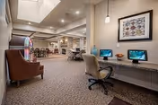 Interior view of a senior living facility common area with a computer workstation on the right, featuring two desktop computers and office chairs. The space has carpeted floors, recessed lighting, and a framed artwork on the wall. In the background, there are tables and chairs arranged for dining or social activities, and a jukebox is visible on the left side.