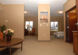 Interior view of a senior living facility room with beige walls and carpet. A round wooden table with a flower arrangement is on the left. In the background, there is a desk with a chair and a lamp near a window, and a bed is visible through an open doorway on the right. A framed picture hangs on the wall in the center.