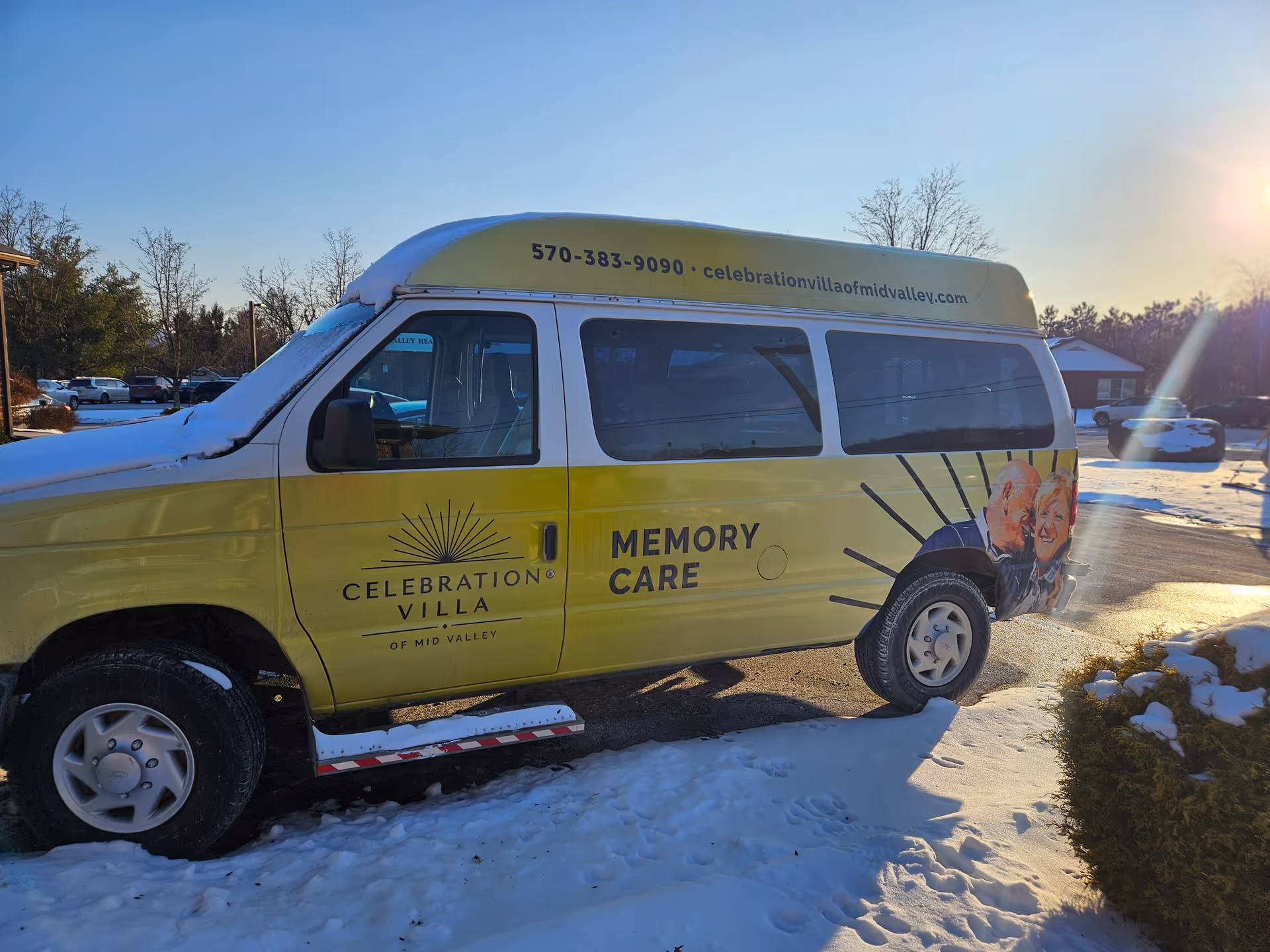 A yellow and white van parked outside on a snowy ground with the sun shining. The van has signage for Celebration Villa of Mid Valley, including their phone number and website, and the words 'Memory Care'. There is an image of an elderly couple smiling on the side of the van.