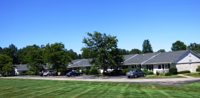 Single-story residential buildings with gray roofs and white walls surrounded by green trees and a well-maintained grassy lawn under a clear blue sky. Several cars are parked along the driveway in front of the buildings.