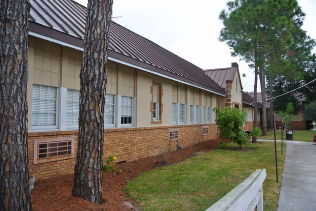 Exterior view of a single-story brick building with multiple windows, a brown metal roof, and a small landscaped area with grass, trees, and a bench in front.