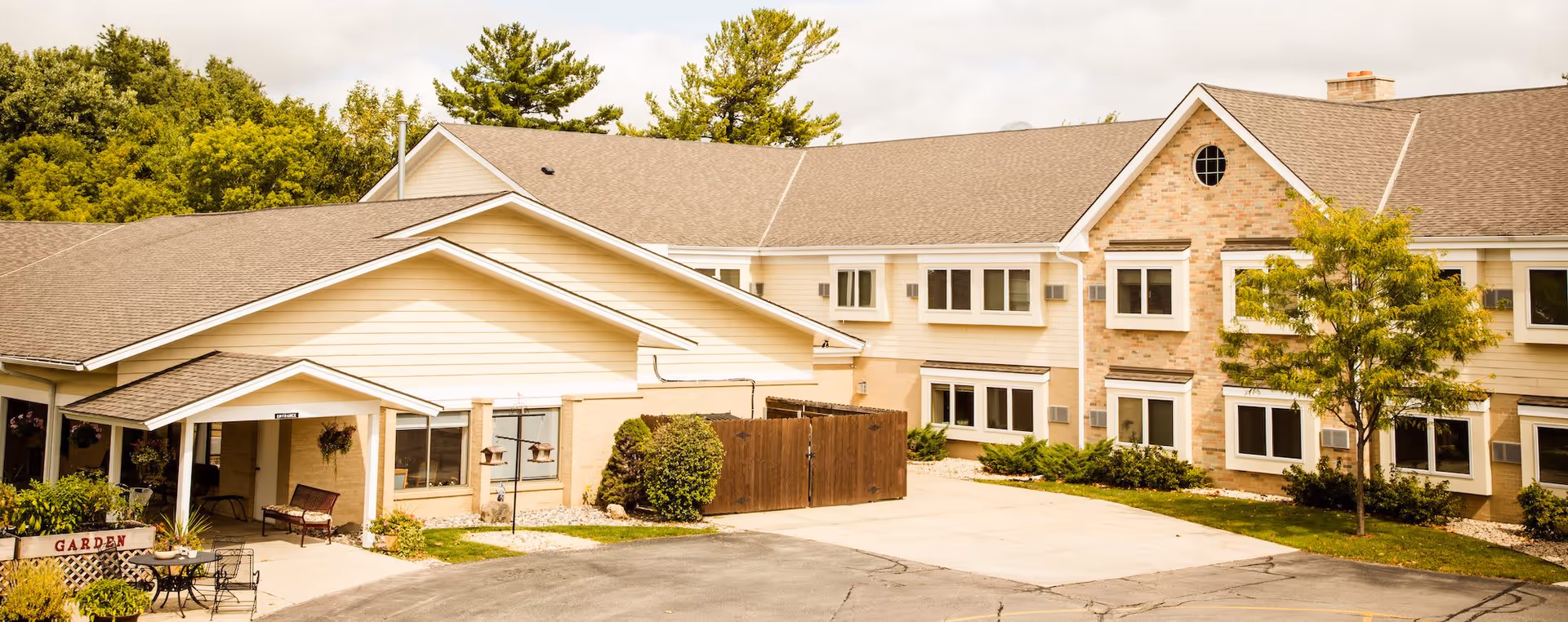 Exterior view of a senior living facility building with beige siding and brick accents, surrounded by greenery and trees. There is a small covered entrance with a bench and hanging plants, a garden sign, and a paved driveway in front.