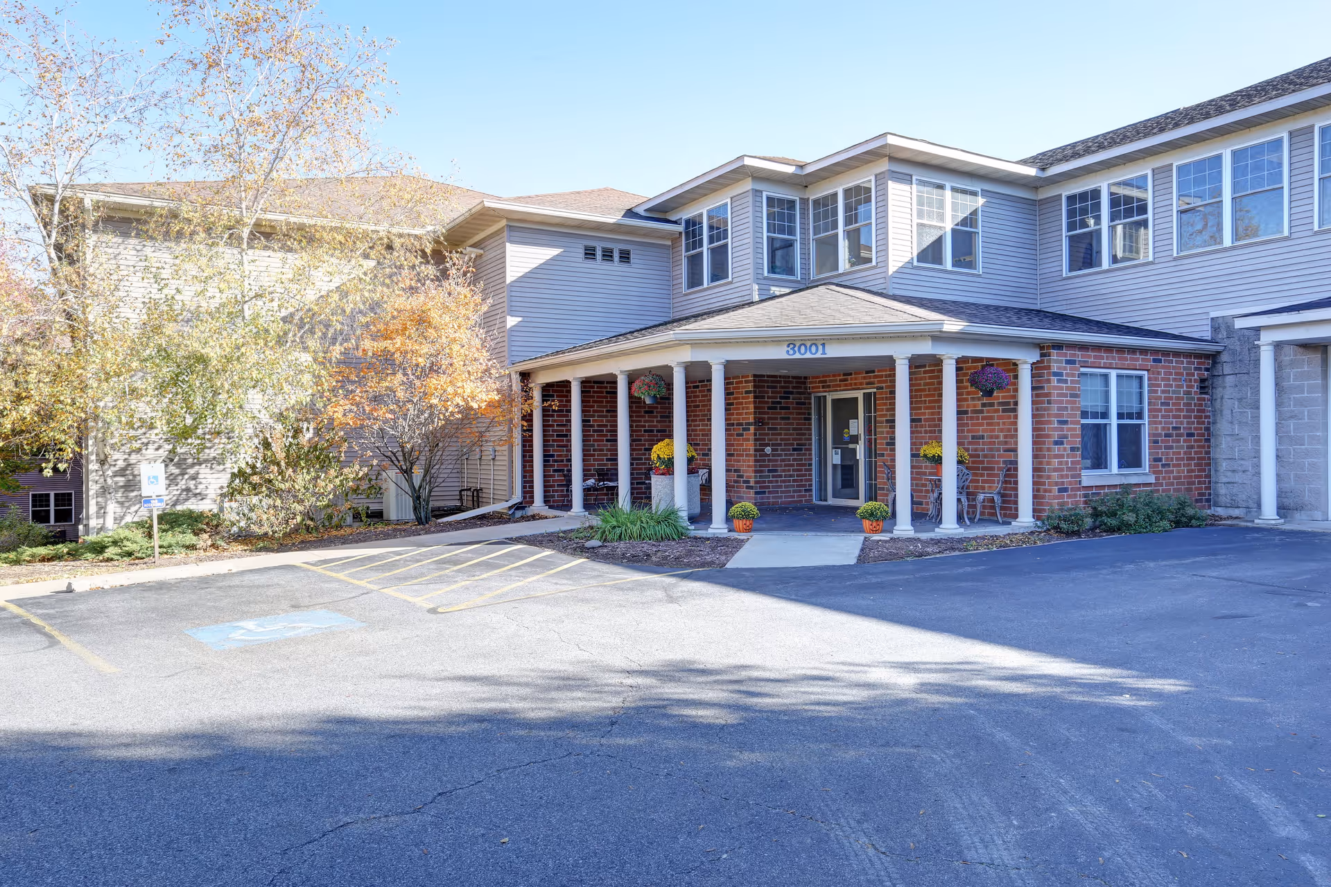 Exterior view of a two-story senior living facility building with a covered entrance supported by white columns. The building has a combination of brick and siding exterior with multiple windows. There are potted plants and hanging flower baskets near the entrance. The parking area in front includes a handicapped parking space and a ramp leading to the entrance. Trees with autumn foliage are visible on the left side.