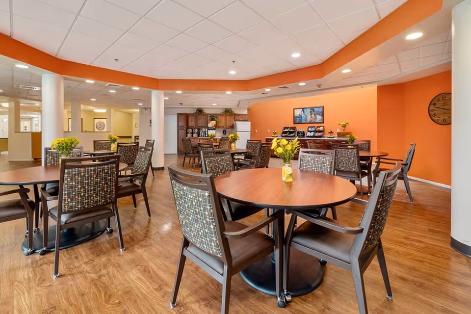 A bright dining area with round wooden tables and patterned chairs, each table decorated with a small vase of yellow flowers. The room has wood flooring, white ceiling tiles with recessed lighting, and an orange accent wall. In the background, there is a kitchen area with wooden cabinets, a refrigerator, and a counter with food trays and decorative items.