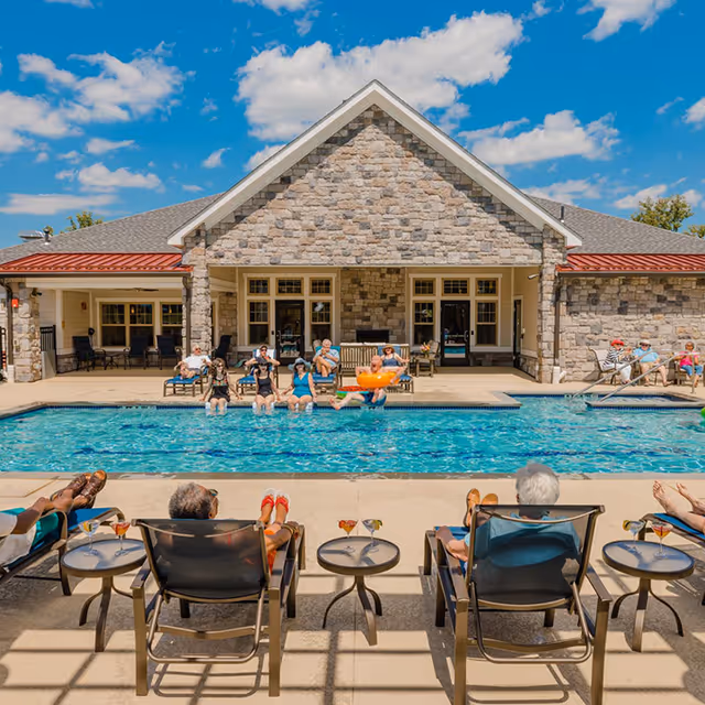 Residents relaxing by an outdoor swimming pool in front of a stone-faced senior living building under a blue sky.