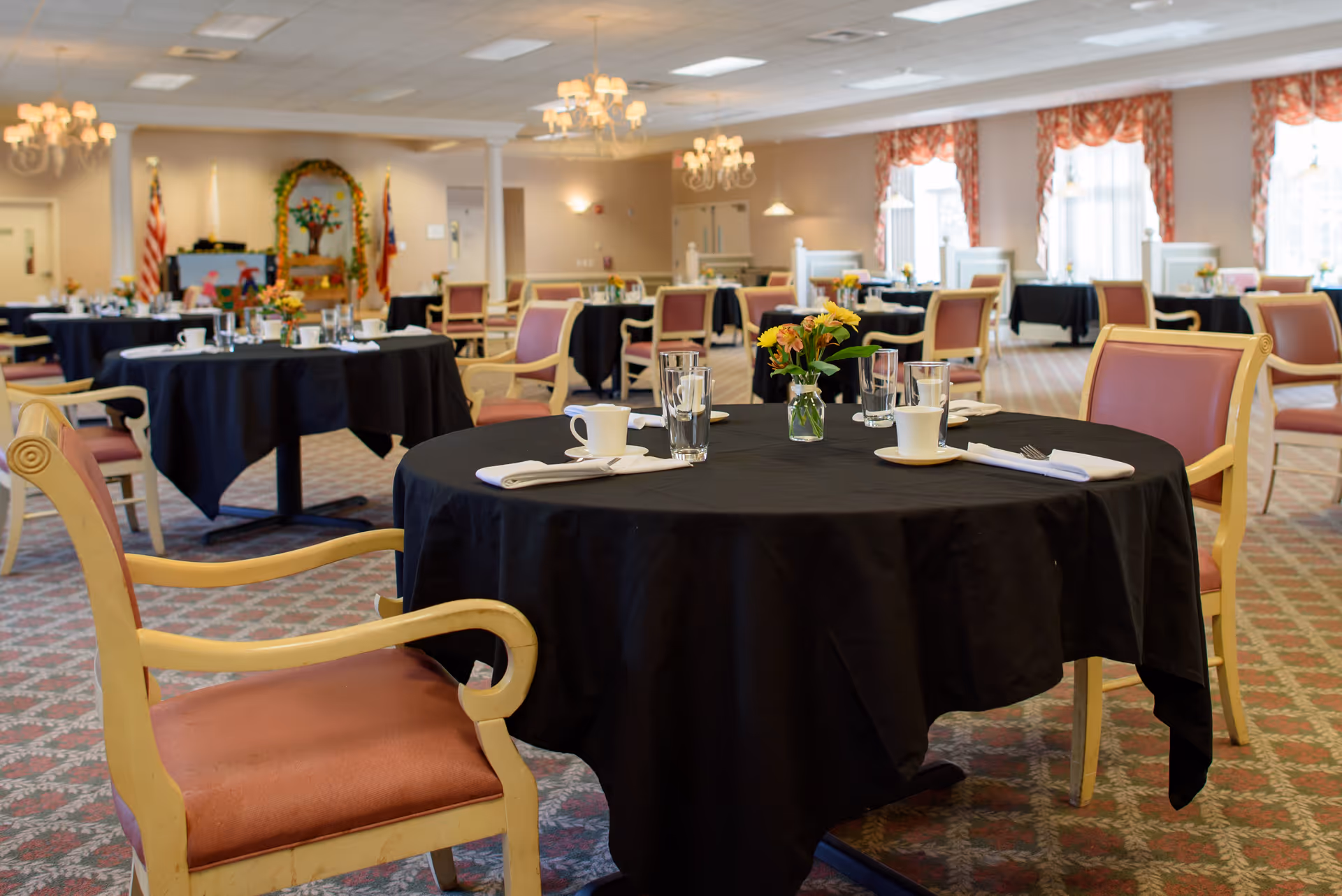 A dining room with round tables covered in black tablecloths, each set with cups, glasses, napkins, and small flower arrangements. The room has upholstered chairs with wooden frames, chandeliers hanging from the ceiling, and large windows with patterned curtains letting in natural light.