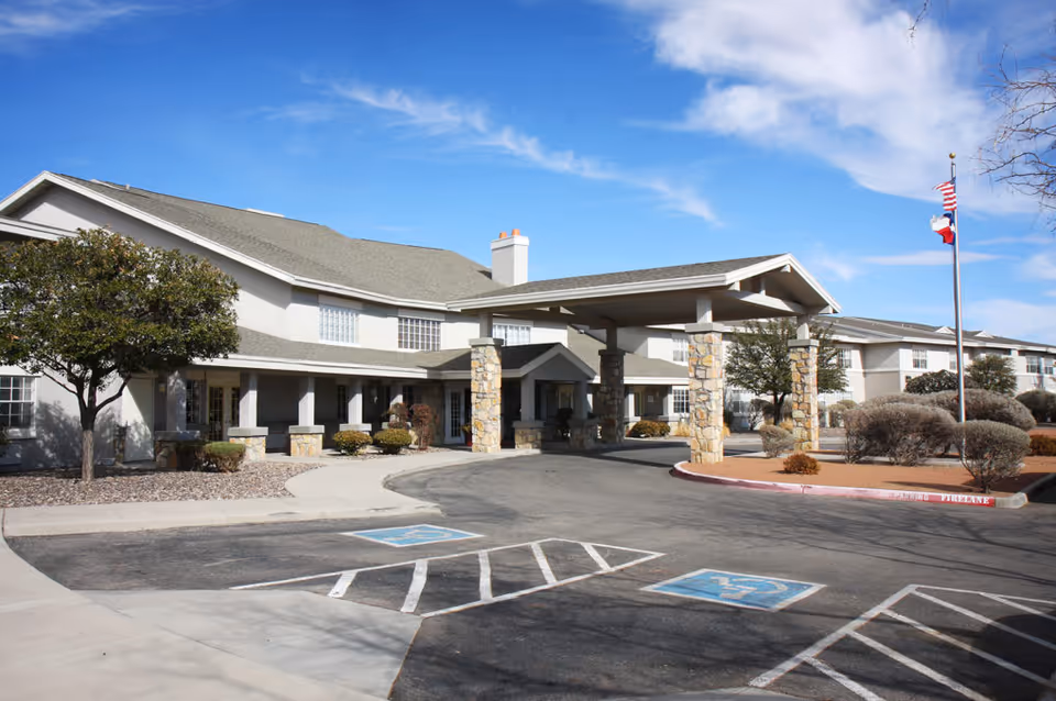 Front entrance of a senior living building with a covered porte-cochere, flagpole, landscaped beds, and accessible parking spaces.