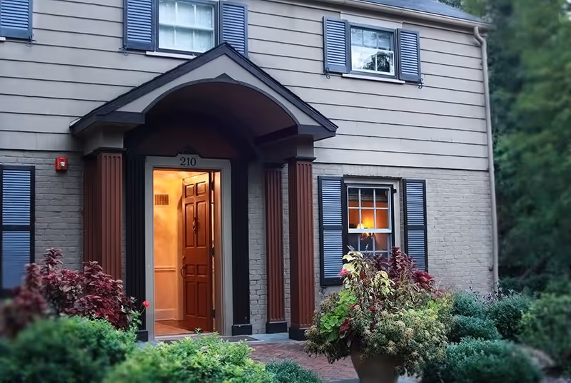 Front entrance of a residential building with the door open, showing warm interior lighting. The building has beige siding, blue shutters on the windows, and a small porch with columns. There are various green and red plants and shrubs in front of the entrance.