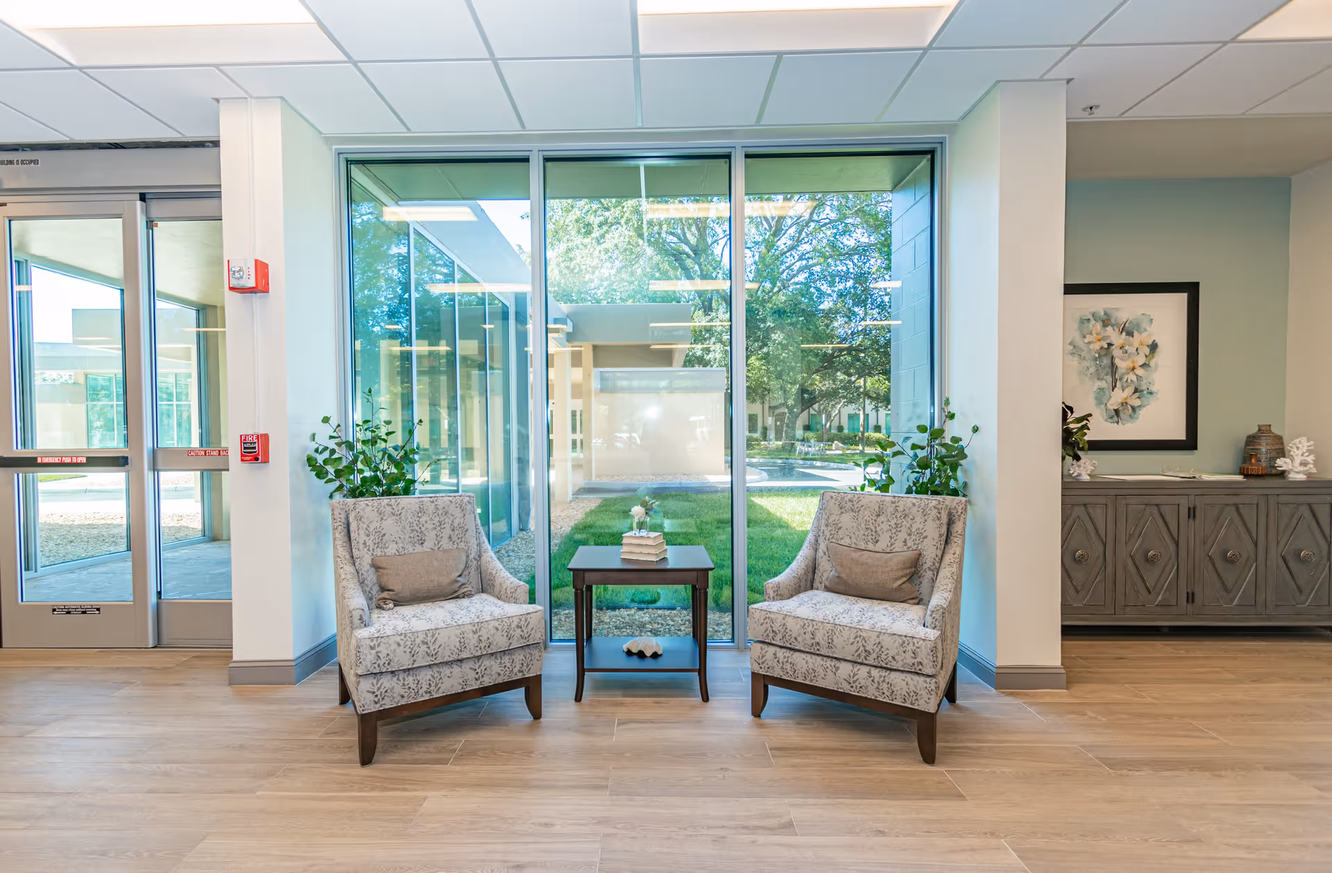 Bright seating area with two patterned armchairs flanking a small table in front of large windows overlooking a courtyard.