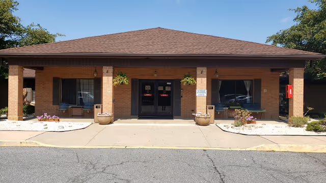 Front exterior view of a single-story brick building with a brown shingled roof, two large windows with black shutters on either side of double glass entrance doors, potted plants and flowers near the entrance, and a clear blue sky above.