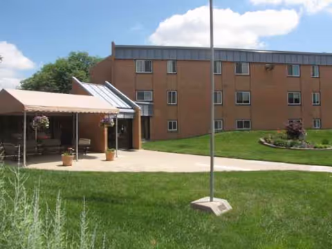 Brick senior living building with a covered patio, green lawn, and walkway.