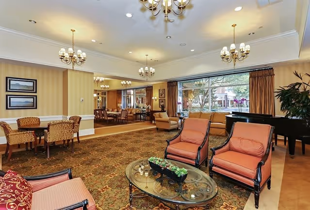 A spacious and well-lit senior living facility common area with comfortable seating including armchairs and sofas arranged around a glass coffee table. The room features large windows with brown curtains, several chandeliers hanging from the ceiling, framed artwork on the walls, and a black grand piano near the window. In the background, there is a dining area with tables and chairs.
