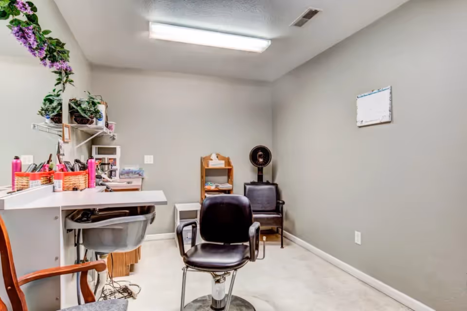 Interior view of a small salon or beauty room with a black salon chair in the center, a sink for washing hair on the left, a large mirror with shelves holding hair products and plants, and a hair dryer chair in the back corner. The walls are painted light gray and the floor is light-colored.