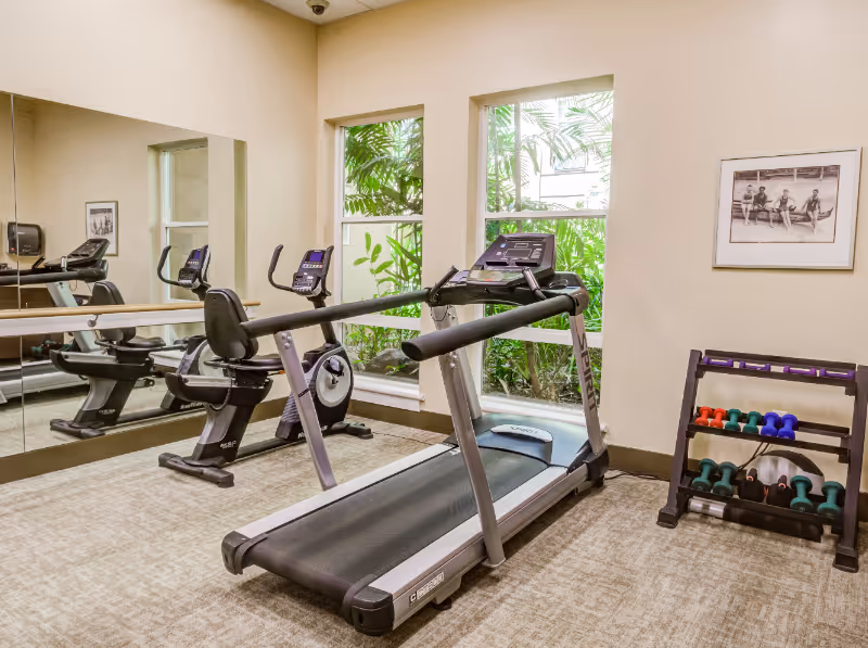 A small fitness room with exercise equipment including a treadmill, two stationary bikes, and a rack of colorful dumbbells. The room has beige walls, two windows with green plants visible outside, a large mirror on one wall, and a framed black and white photo on another wall.