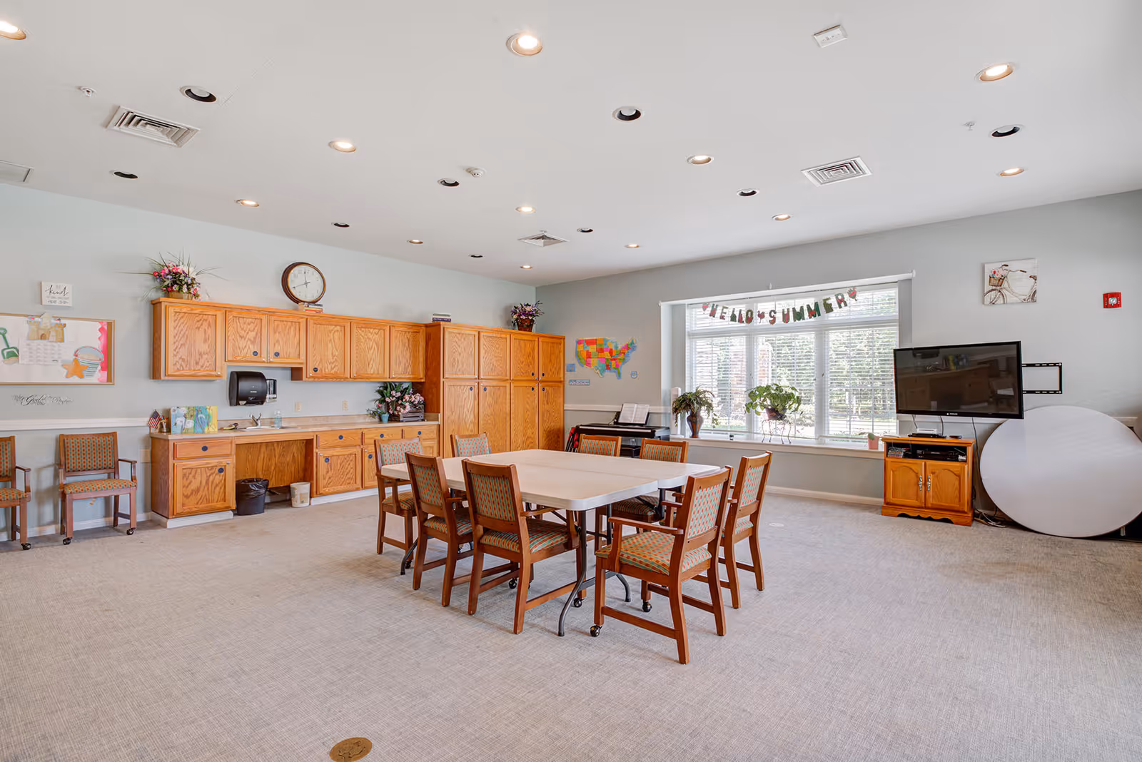 A bright and spacious common room with a large window decorated with a 'Hello Summer' banner. The room features a rectangular table surrounded by eight wooden chairs with patterned cushions. Along one wall are wooden cabinets and a countertop with a clock above, a paper towel dispenser, and some decorative plants. There is a TV mounted on the wall with a wooden stand below it, and a piano is visible near the window. The room has recessed lighting and a light-colored carpeted floor.