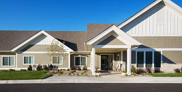 Front exterior view of a single-story senior living facility building with beige siding, white trim, a covered entrance with two chairs, and a well-maintained lawn and landscaping under a clear blue sky.