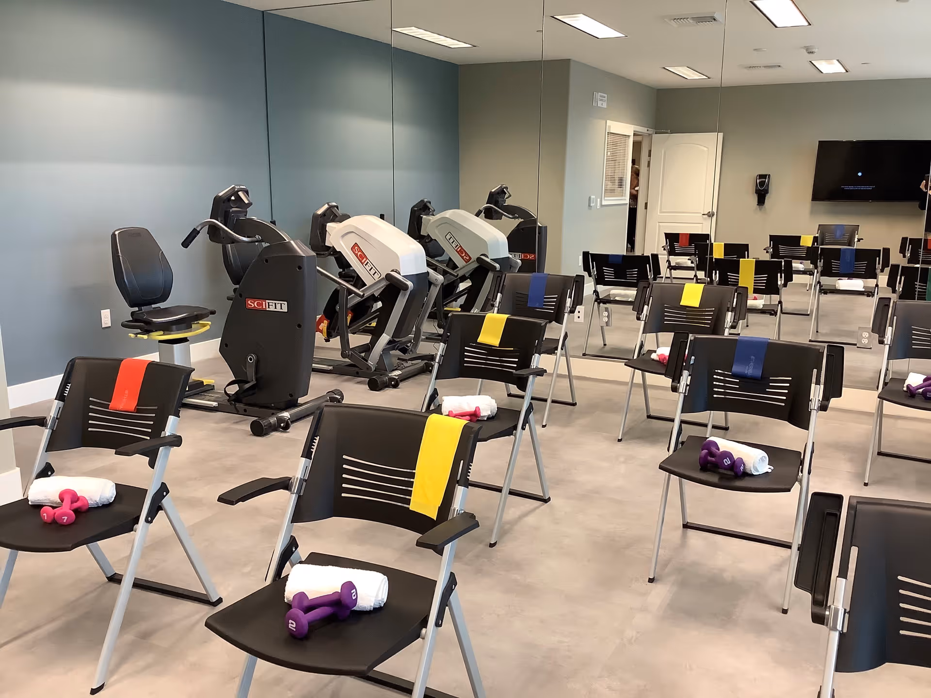 Exercise room with multiple black chairs arranged in rows, each chair holding a rolled white towel and a pair of small dumbbells in various colors. In the background, there are exercise machines against a wall with large mirrors reflecting the room. The walls are painted in a muted blue and gray color, and a flat screen TV is mounted on the far wall.