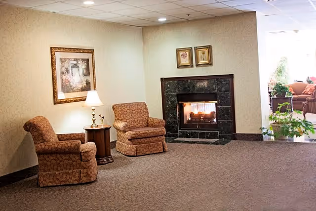 A cozy sitting area with two patterned armchairs and a small round wooden table with a lamp between them. There is a framed painting on the wall above the lamp. To the right, there is a black marble fireplace with a fire burning inside. In the background, another seating area with sofas and plants is visible.