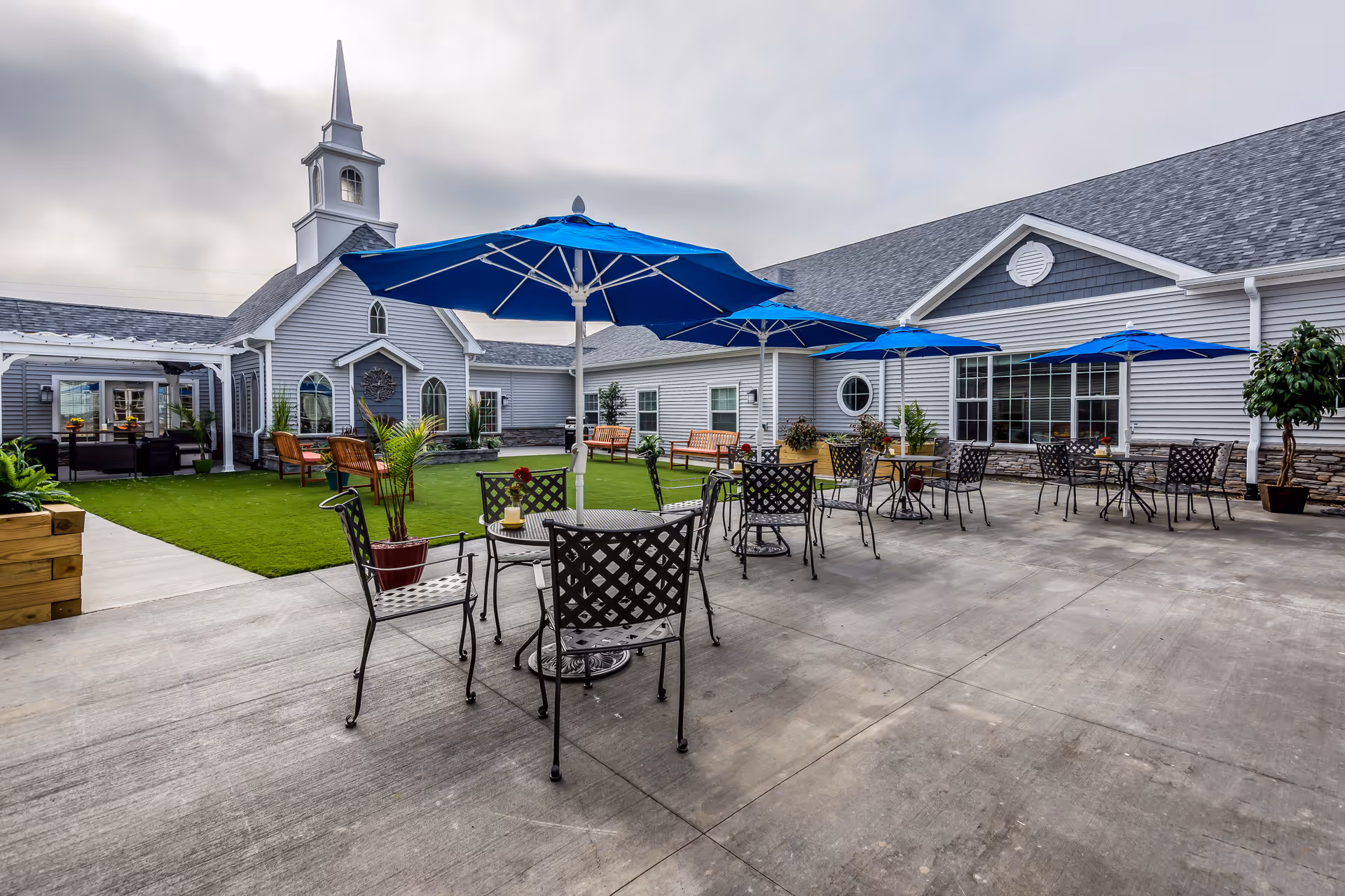 Outdoor courtyard patio with metal tables, blue umbrellas, seating and a lawn in front of a light-gray senior living building with a steeple.