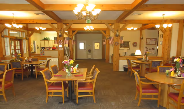 Interior view of a dining area in an assisted living facility with multiple wooden tables and chairs arranged neatly. Each table has a small flower vase and condiments. The room features wooden beams on the ceiling, chandeliers, and an open kitchen area in the background. American flags and wall decorations are visible on the walls.