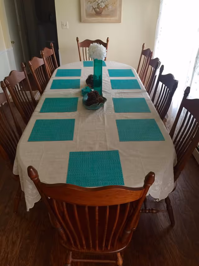 A long dining table set with teal placemats, a white tablecloth, a floral centerpiece, and wooden chairs on both sides.